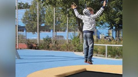 A young boy in a stripey hoodie bounces on a trampoline in an outdoor playground.