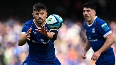 Leinster player Ross Byrne - who has short brown hair and a short beard - reaches to catch a rugby ball flying towards him. He is wearing the dark blue Leinster home kit. Behind him out of focus another Leinster player is jogging past