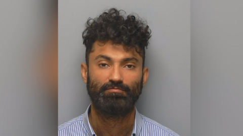 Mugshot of Diwan Khan, who is wearing a striped blue and shirt shirt and has a black beard and black curly hair.