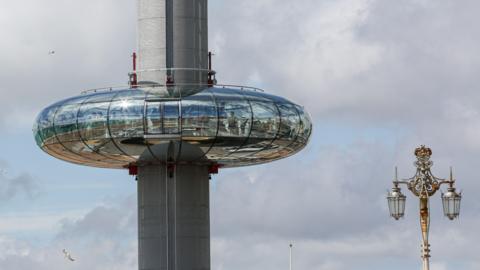 This close up image shows the doughnut-shaped glass viewing capsule of the i360 in mid-air part way up the tower against a cloudy skyline. The top of a street lamp of visible on the right hand side of the photo. 