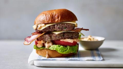 A double-patty burger, topped with cheese, lettuce and tomato sits on a board alongside a bowl of burger sauce