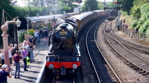 Flying Scotsman locomotive arrives at Bridgnorth. The green loco has a black boiler cover with a sign that reads Severn Valley reopening train. Behind the loco are seven brown carriages with people on the platform taking photographs with their mobile phones
