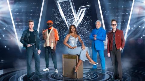 The Voice coaches in front of a silver background with a big letter 'V'.