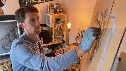 Matt Sayers, dressed in a blue short and wearing a latex glove as he draws with charcoal at an easel in his workshop by a bare lightbulb