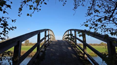 Bright blue cloudless skies lie on the other side of a curving bridge over a river. The bridge is framed by branches of trees with green fields just visible on the other bank.