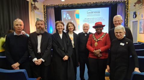 A group of eight people stood in a line smiling at the camera inside Lucem House Community Cinema during its 10 year anniversary