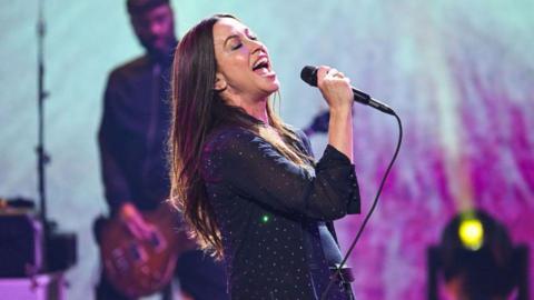 Alanis Morissette performing at Dick Clark's New Year's Rockin' Eve in black shirt with sequins. She is holding a microphone and is singing with eyes closed