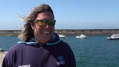 Dan Slimm is smiling at the camera as he stands on the quayside on a bright sunny day. He has shoulder-length curly hair and is wearing sunglasses. His hoodie is branded and reads SKIPPER ALDERNEY FERRY SERVICES. Behind him are four small boats moored inside the harbour.