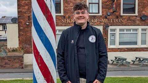 A smiling man stood to next to a maypole, painted red, white and blue in an outdoor setting. In the background is a pub called The Shoulder of Mutton. The man has short, brown hair and is wearing glasses, a long blue jacket and beige trousers. 