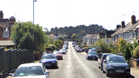 A residential street is shown with cars lining both sides of the road in the centre, with fences and trees in front of some homes and a woodland area seen in the background at the end of the road.