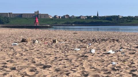 The sandy South Shields beach with litter strewn across it in the foreground. The red Herd Groyne Lighthouse can be seen on a rocky outcrop into the sea in the background. Behind that is the southern skyline of Tynemouth.