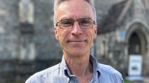 Dr Murrison smiles at the camera - he is wearing glasses and a blue shirt. The background is blurred, but it is a sunny day in front of a church