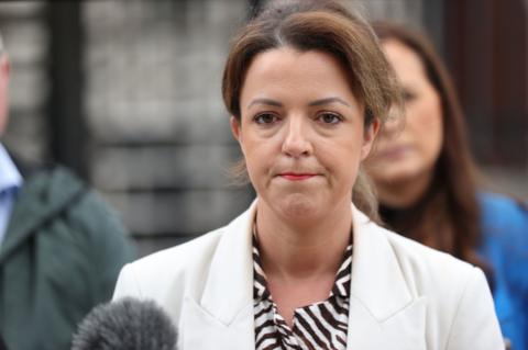 A woman in a white jacket and black and white blouse, with short brown hair, is about to speak into a microphone. She is outside a building. In the background, other people can be seen but they are out of focus