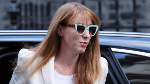Angela Rayner exiting a car wearing mint green and tortoiseshell glasses, a white jacket and top