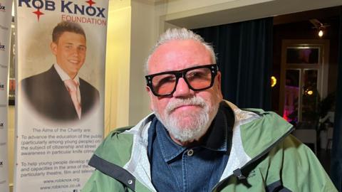 Actor Ray Winstone is wearing a denim shirt and green jacket with tinted glasses. Behind him is a poster that says "The  Rob Knox Foundation" and has a picture of a young man in a suit and pink tie.