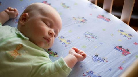 A photo of a baby sleeping on its back in a cot. The baby is wearing a pale green sleepsuit. The bed sheet has stars and vehicles on it.