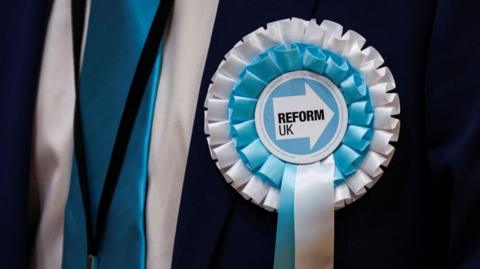 A close up picture of a Reform UK blue and white rosette attached to a suit.