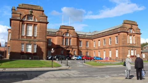 A sandstone council headquarters in Dumfries on a sunny day with cars parked in front and a blue sky above