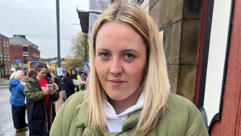 Hannah Collins has blonde hair and is wearing a green puffa jacket.