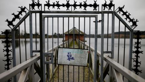 A sign hangs on a metal gate at Severn Trent Water's Cropston Reservoir. A bridge can be seen leading towards a brick building.