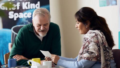 A woman dressed in a shawl sits at a table with a man in a green jumper. She is showing him what appears to be a physical photograph. Some coffee mugs and a box of tissues sit on the table in front of them.