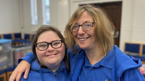 Two women smiling and looking straight at the camera with their arms around each others shoulders. They are both wearing blue polo shirts and jumpers.