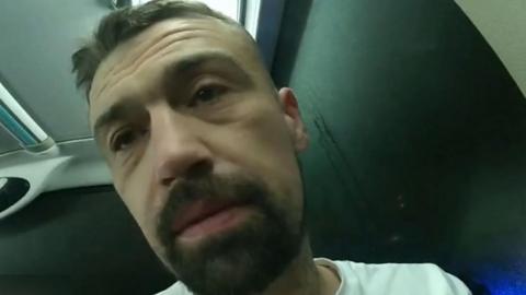 A man with a brown beard and wearing a white T-shirt while on a bus