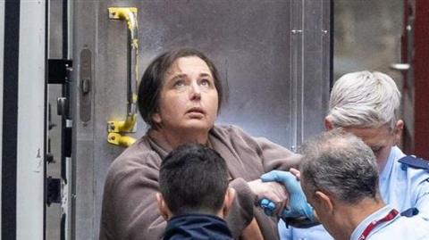 Erin Patterson appears handcuffed as she is escorted out of a custody van. She is wearing a brown coat and her black hair appears to be tied back, She is looking upwards. 
There are three custody officers - two of them have their back turned to the camera. The third's face is obscured by one of the other custody officers. 
It appears that she is being taken off the van and lead into court building. 