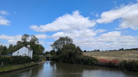 A waterway dominates the picture with a grassy bank and trees on the right-hand side and trees and a white-walled house in the left-hand side. It is a sunny day with white clouds in the sky