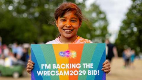 Rose, merch ysgol gwallt brown, yn dal cerdyn gyda neges yn cefnogi cais Wrecsam2029