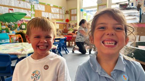 The image shows two Year One pupils sat with big teethy grins showing their teeth covered in toothpaste. The picture is taken in a class room and the children have both got stickers on their school uniforms. 