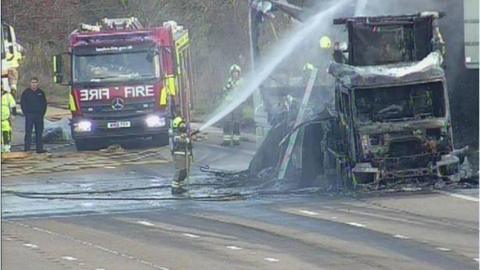 A firefighter using a hose on a lorry that is burnt out. A fire engine is on the road and other firefighters are watching on. There is oil and water on the road.