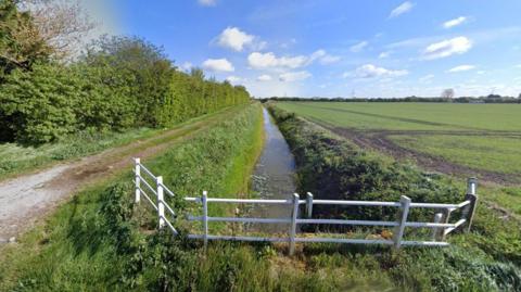 A ditch in the Fens which is partly filled with water. There is a white fence straddling the ditch which is bordered b a farm field on one side and a bridleway on the other. A blue sky in the background and hedgerow on the left side of the path.