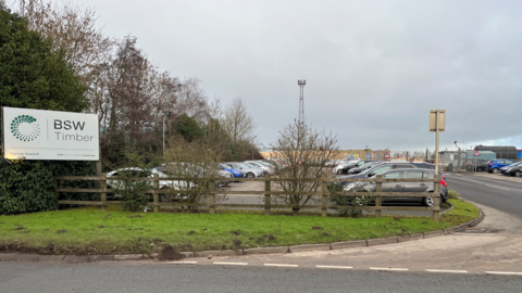 A car park sits next to a main road. There is BSW Timber signage to the left of the car park. 