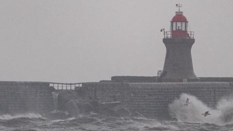 Damage is visible to the pier structure which has large rocks in front of it. There is a light house, large waves and three birds in flight.