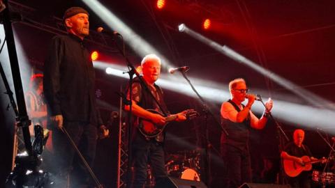 Three older men performing on a dark stage that is lit with a few red lights and white spotlights. They are all standing behind microphones.