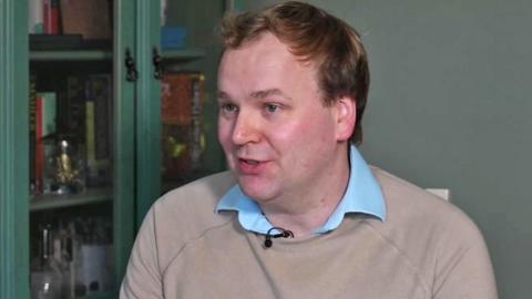 William Wragg, former MP, speaking to the BBC's Joe Pike in his home. He is wearing a blue open necked shirt and light brown jumper and sitting in front of a cabinet 