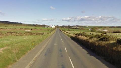 Part of the A2 Whitebridge Road in Onchan, which is a long straight with fields on either side and a large white building in the distance. There are blue skies with some while clouds above.