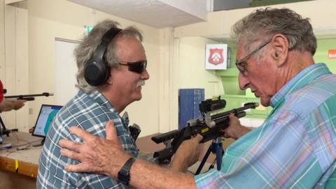 An instructor helps a visually impaired shooter position a competition air rifle during an adapted target shooting session