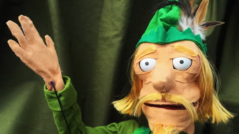 A puppet with its hand raised on the left. It is a man with a green hat and it has blonde hair and a moustache. It is against a green background.