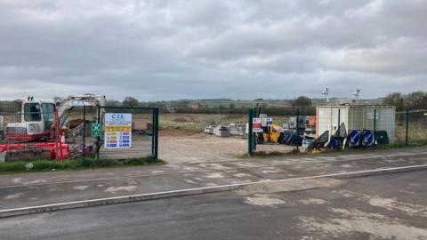 Picture shows fenced land with green scenery in the background and from within the fences there are road signs, a storage unit and a digger.