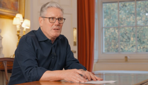 Sir Keir Starmer is seated at a polished wooden table in a well-lit room, wearing a dark long-sleeved shirt, with hands resting on a letter to his son. Behind him is a large window with autumn foliage visible outside, flanked by orange curtains.