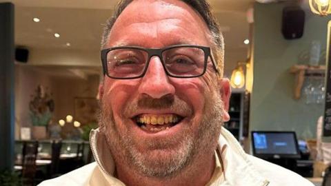 Malcolm Barry, a man with salt and pepper couloured hair and a light beard, grins at the camera. He is wearing black-framed glasses.