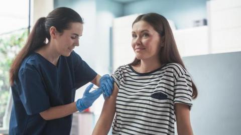A woman is getting a flu vaccine from a nurse who is wearing a navy uniform and blue gloves. The background is blurred and shows a health clinic with plants outside the window.
