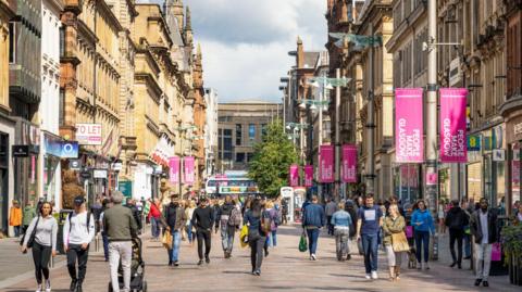 A stock image of Glasgow's Buchanan Street. There are crowds of shoppers walking in both directions. On the lampposts there are banners which say 'People make Glasgow' on them. Multiple shops are visible on the left and the right of the image.