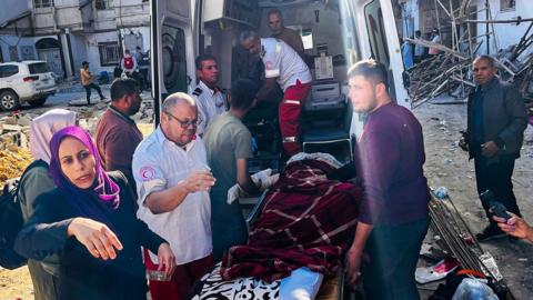 A group of people and medics, including people wearing shirts with the Palestinian Red Crescent logo, lift a wounded patient on a stretcher into the back of an ambulance on a street, with damage and debris in the background, outside the Kamal Adwan Hospital in northern Gaza on 28 October