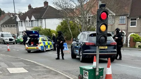 Armed police stood next to a police car with a traffic light in front of them
