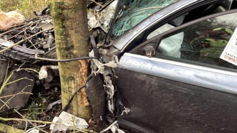 The front of a car smashed up with a tree against its front passenger side. Bits of the car are scattered around the trunk and wires trail from the car.