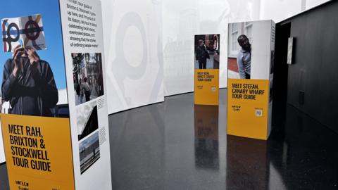 An exhibition space with tall boxes in a room. The boxes have pictures and text, telling the stories of the tour guides working for Unseen Tours. 