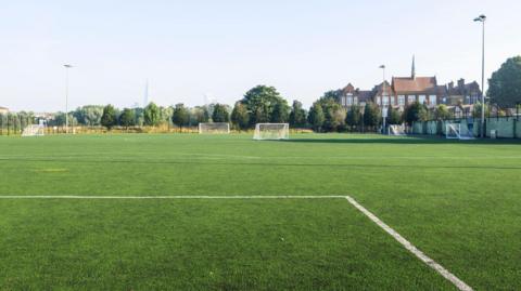 The image shows an empty leisure field in a residential district, with a view of the city in the distance. The sky is cloudless and light blue. The white lines of a football pitch can be seen in the foreground of the image.